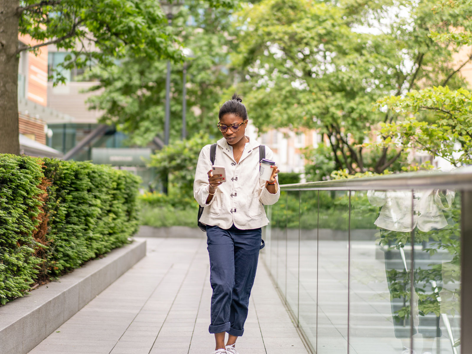 Une étudiante qui marche à l’extérieur sur le campus d’une école, tenant une tasse de café dans une main et regardant son téléphone portable.