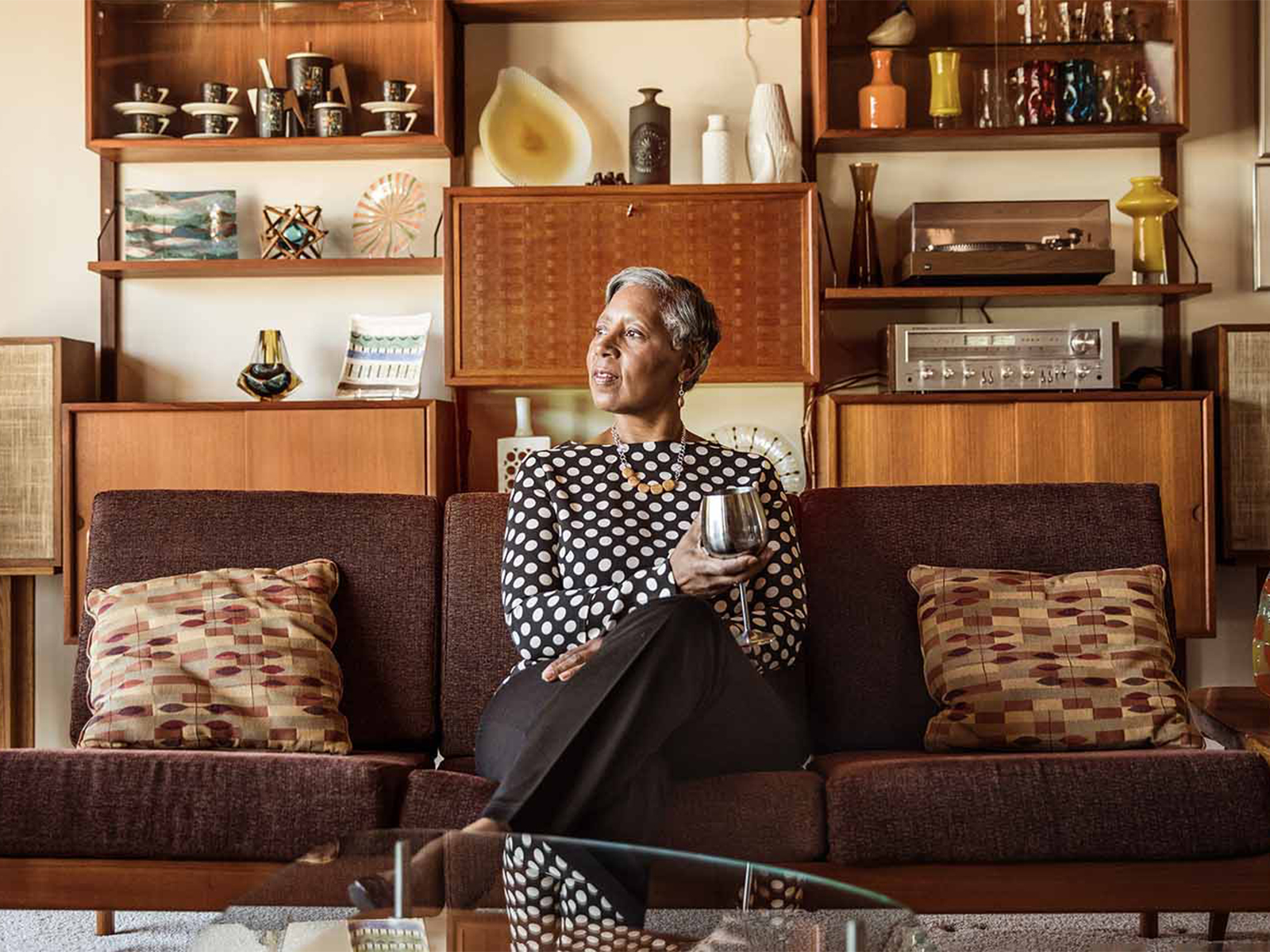 An older woman sitting on a couch, with a stemmed silver cup in her hand, looking out the window.