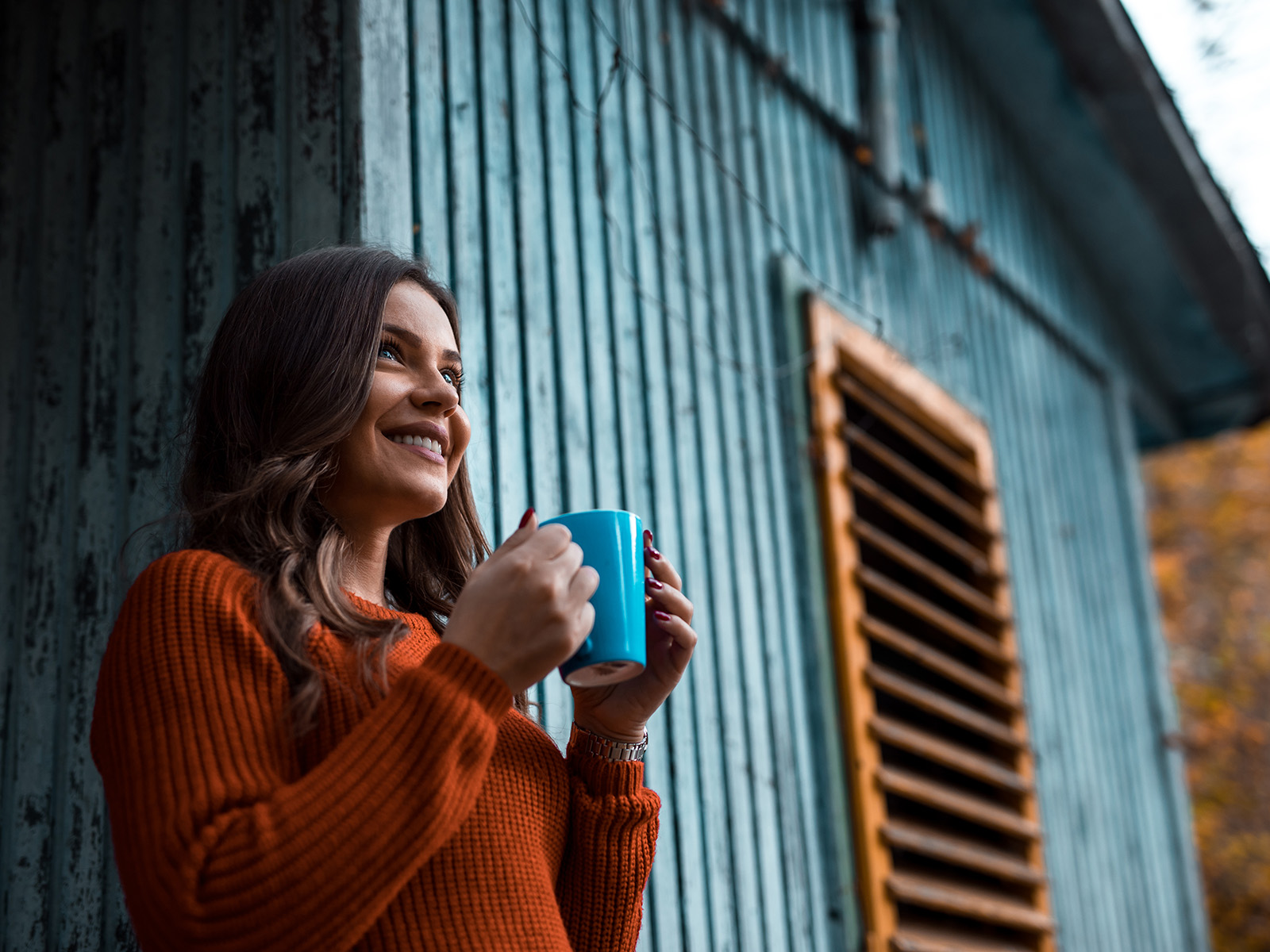 Smiling, relaxed-looking woman holding a coffee mug, standing next to a blue wooden house.