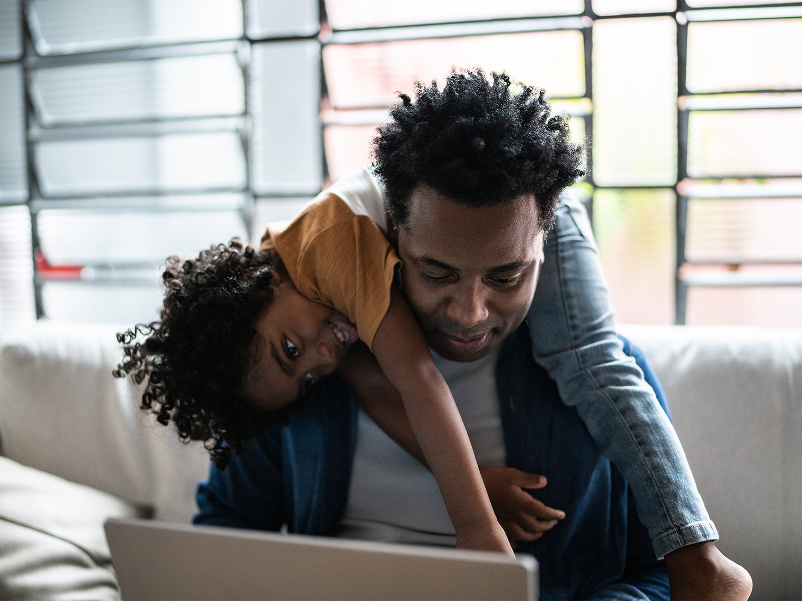 A father looking at his laptop with his smiling child laying across his shoulders.