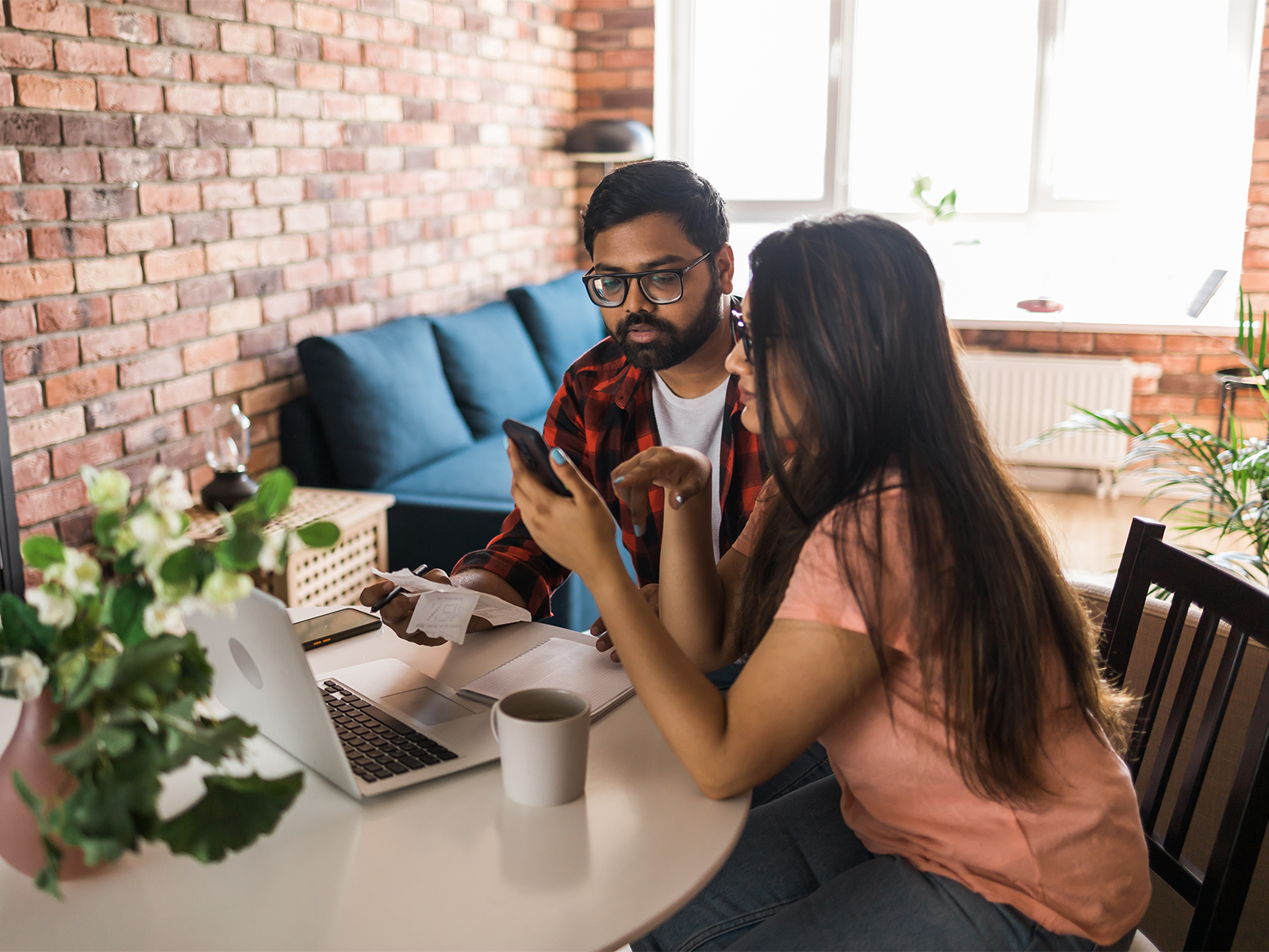 A couple sitting at a dinner table in front of a laptop and receipts, looking at the smartphone in the woman's hand. 