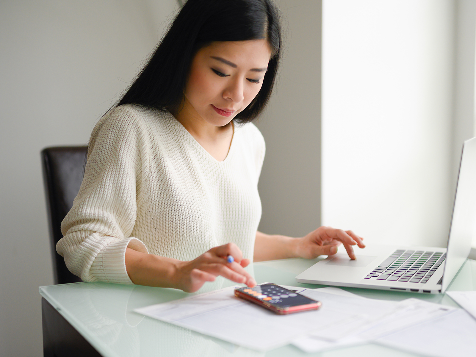 A woman using the calculator on her smart phone to input some numbers on her laptop.