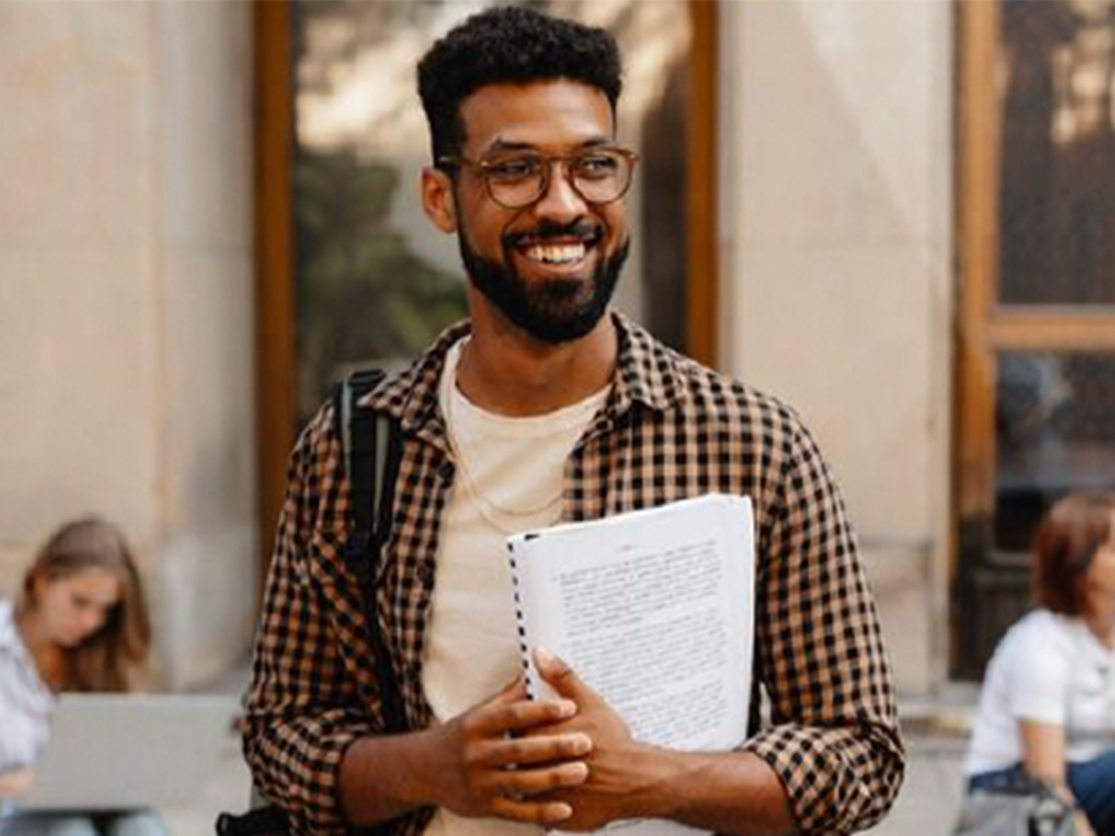 A smiling male student standing outside a building while holding a notebook in his hands.