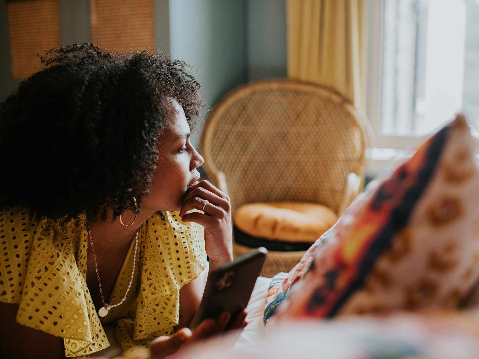 Une femme semble méditer sur la vie, en regardant par la fenêtre de sa chambre. Elle est sur son lit allongée sur le ventre, et elle se touche le menton. Elle tient un téléphone, mais ne le regarde pas.