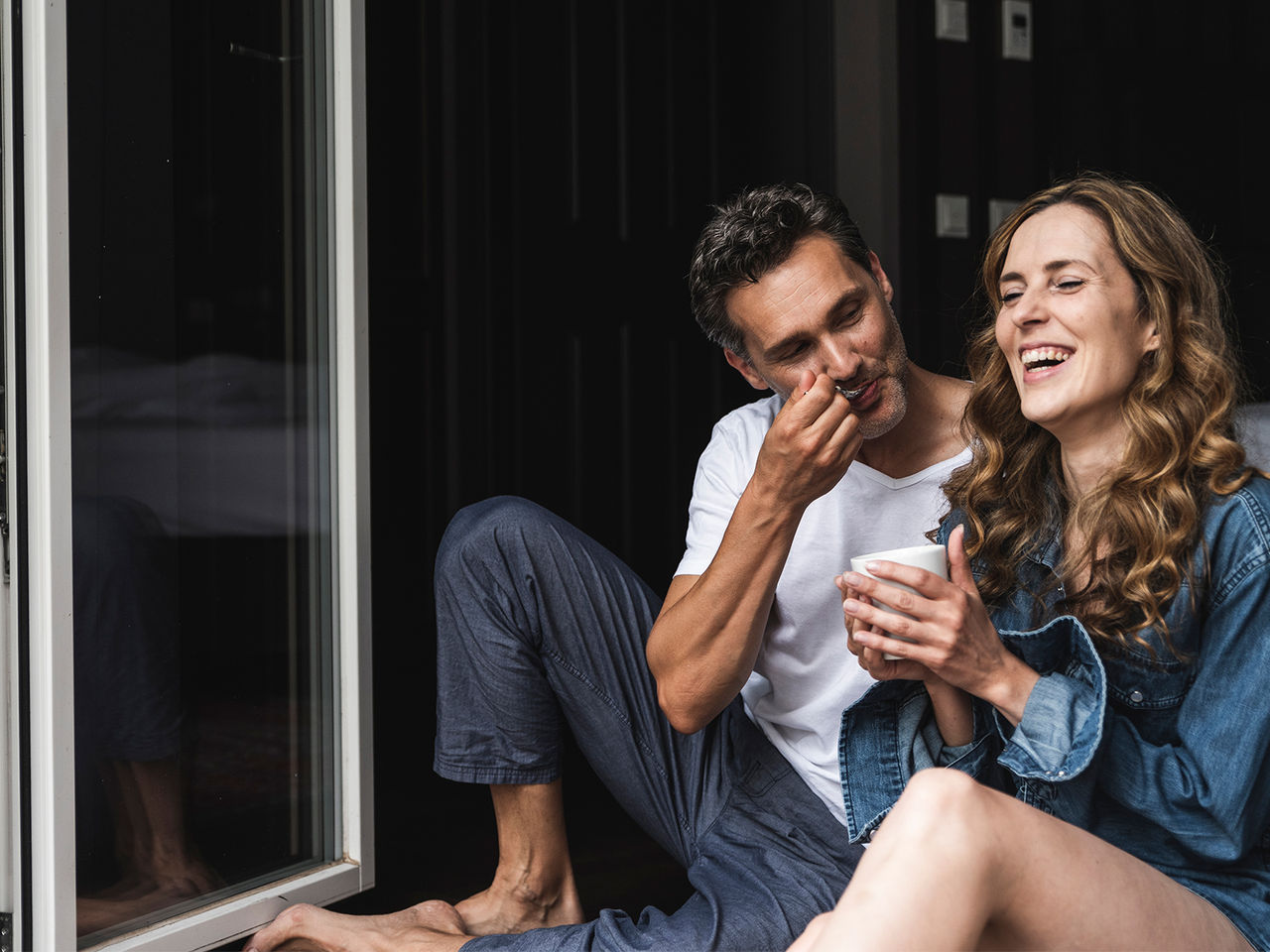 A couple sitting in front of open glass doors, eating breakfast together on the floor of their bedroom.