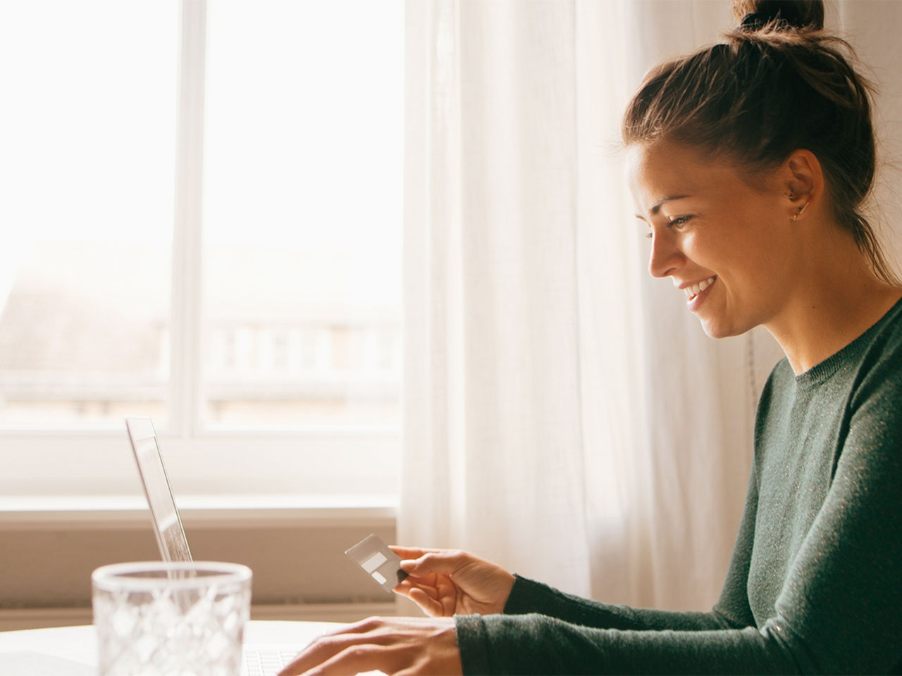 A young woman holding up her credit card and entering its numbers onto her laptop.