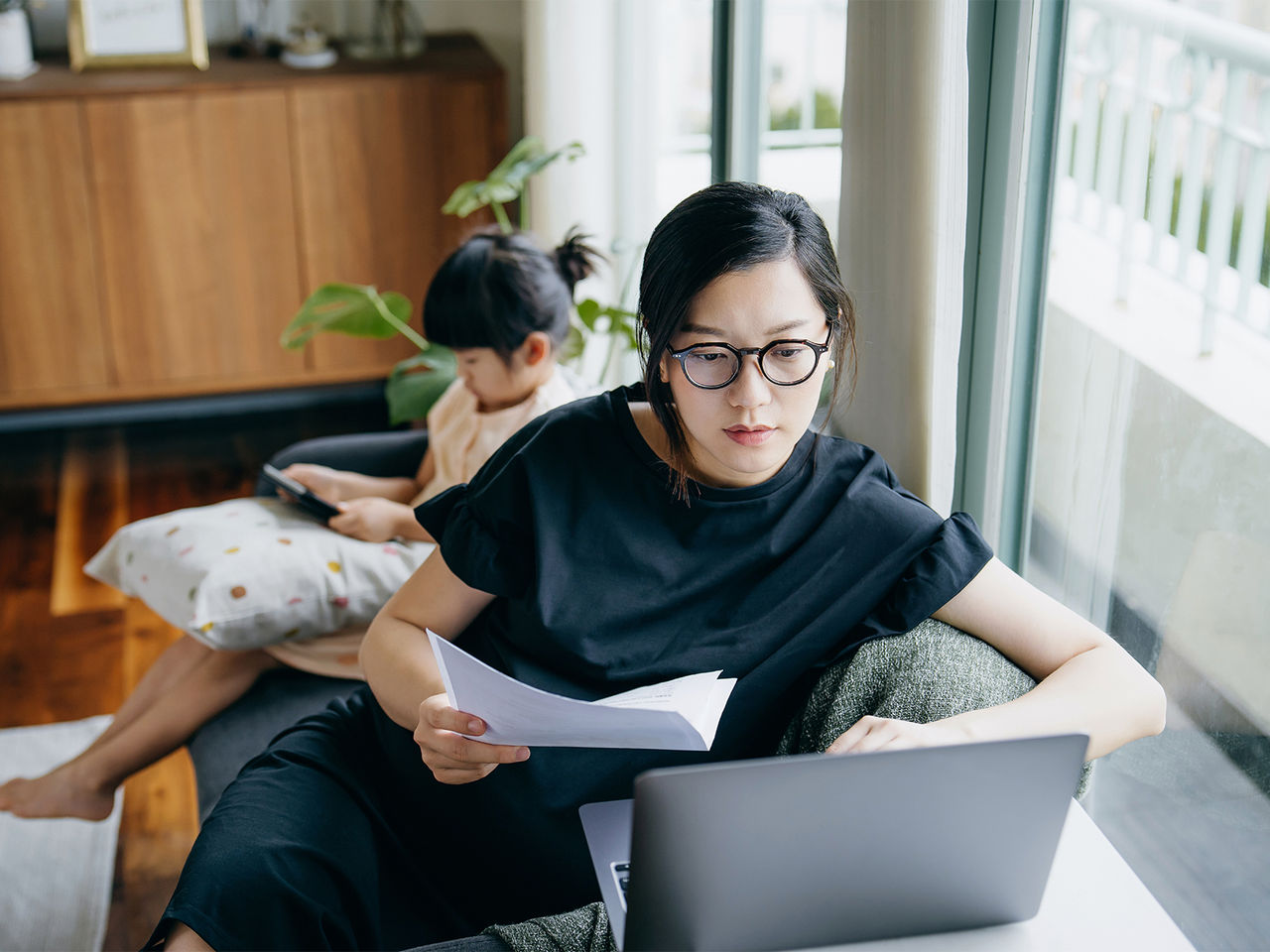 A woman holding documents in her hand and looking at a laptop. Her child is playing on a tablet behind her.