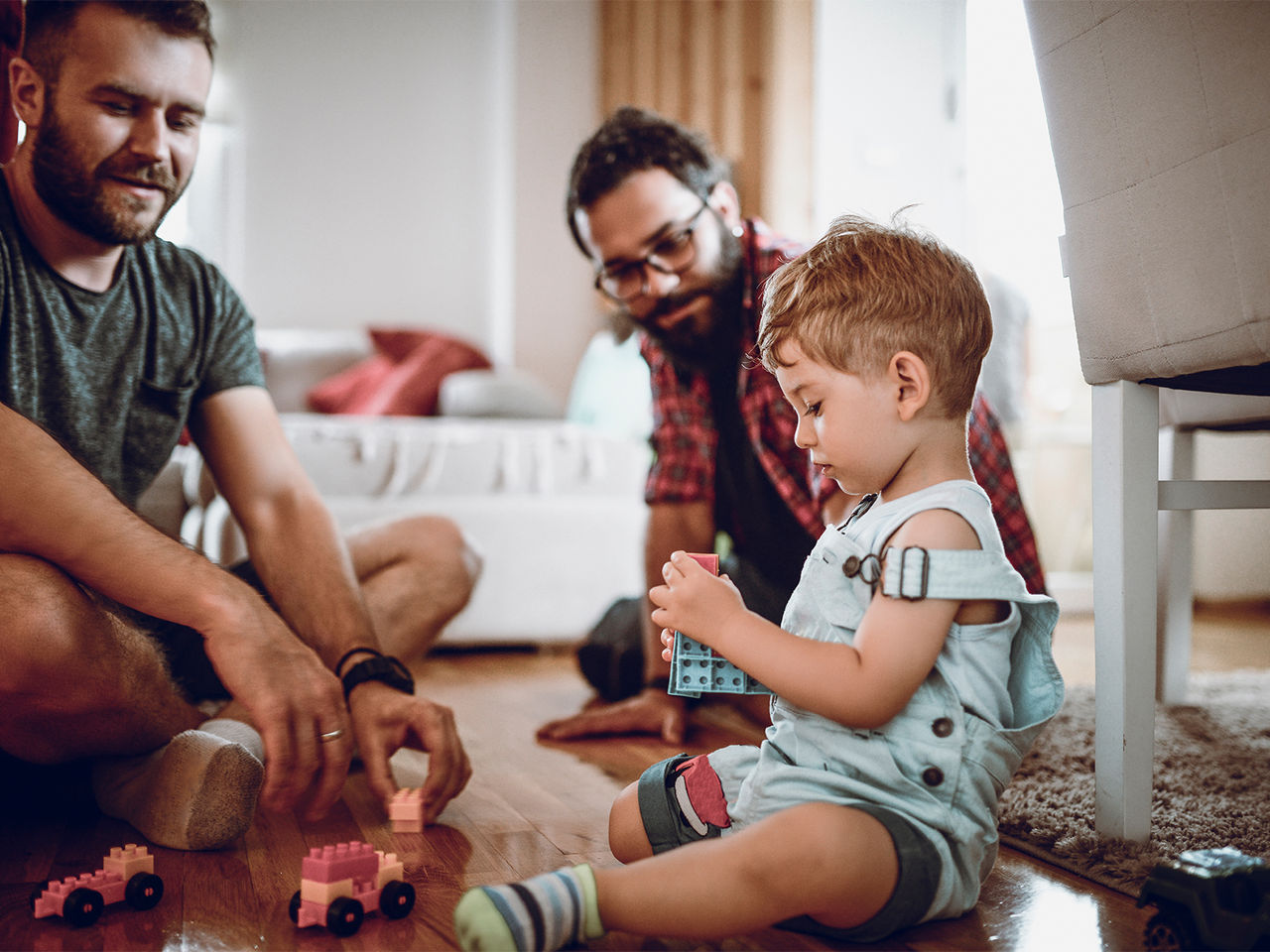 Two fathers and their young child playing with toy building blocks.