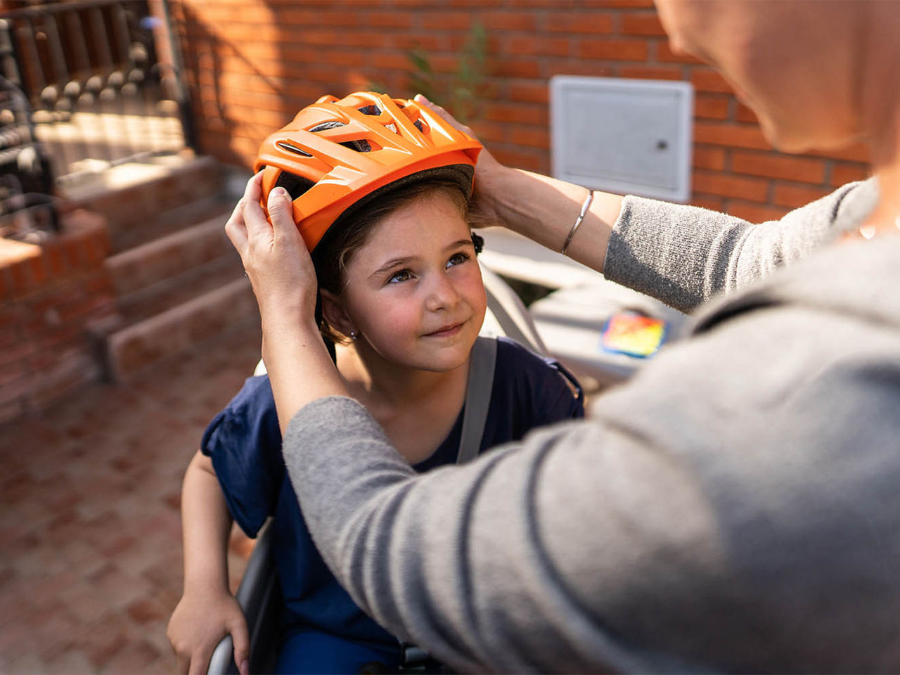 A mother placing an orange bike helmet on her young daughter's head.