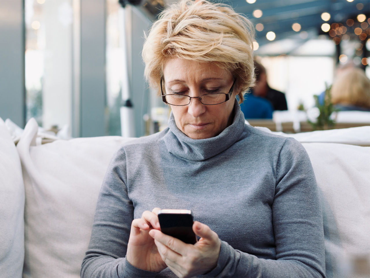 An older woman, sitting on a couch, looking down at the smartphone held between her hands. 