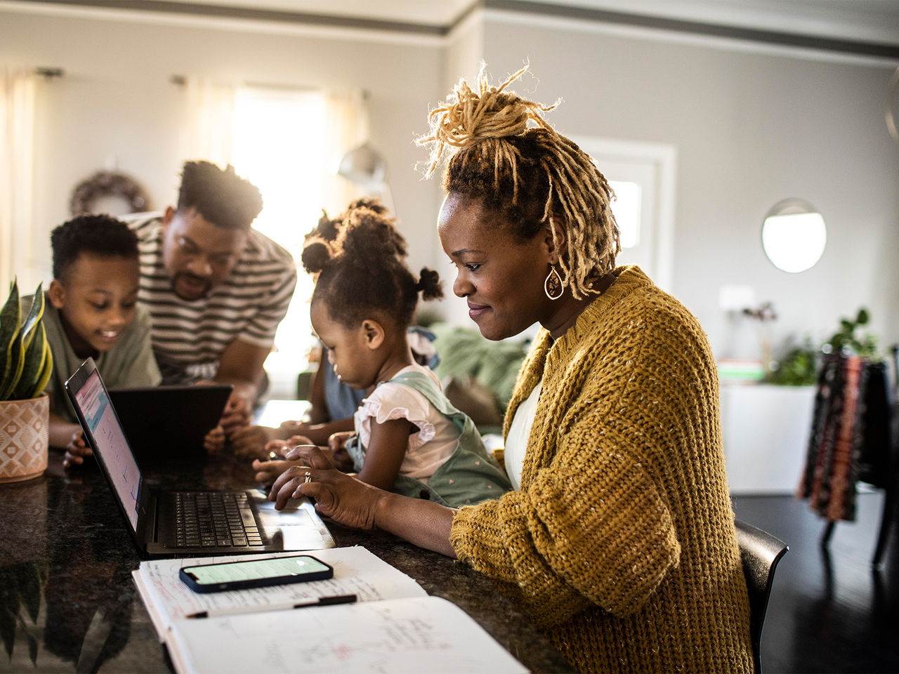 A mother using a laptop in the kitchen, with her budget notebook at her side, while her kids and husband play on a tablet.