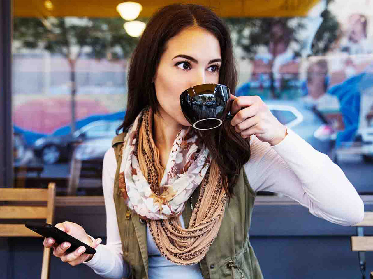 A young woman sitting outside a local coffee shop, sipping on a cup with her smart phone in the other hand.